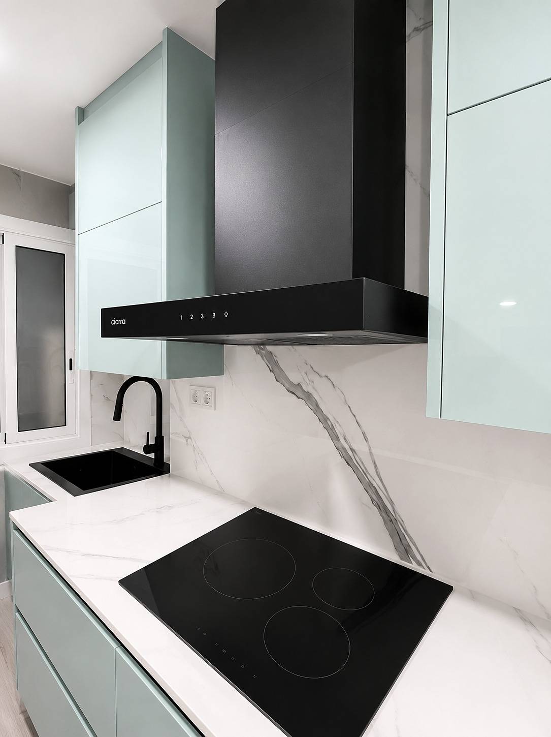 Cocina con muebles altos y bajos verde agua, campana negra sobre la placa de inducción y frontal blanco con veta, ya montada