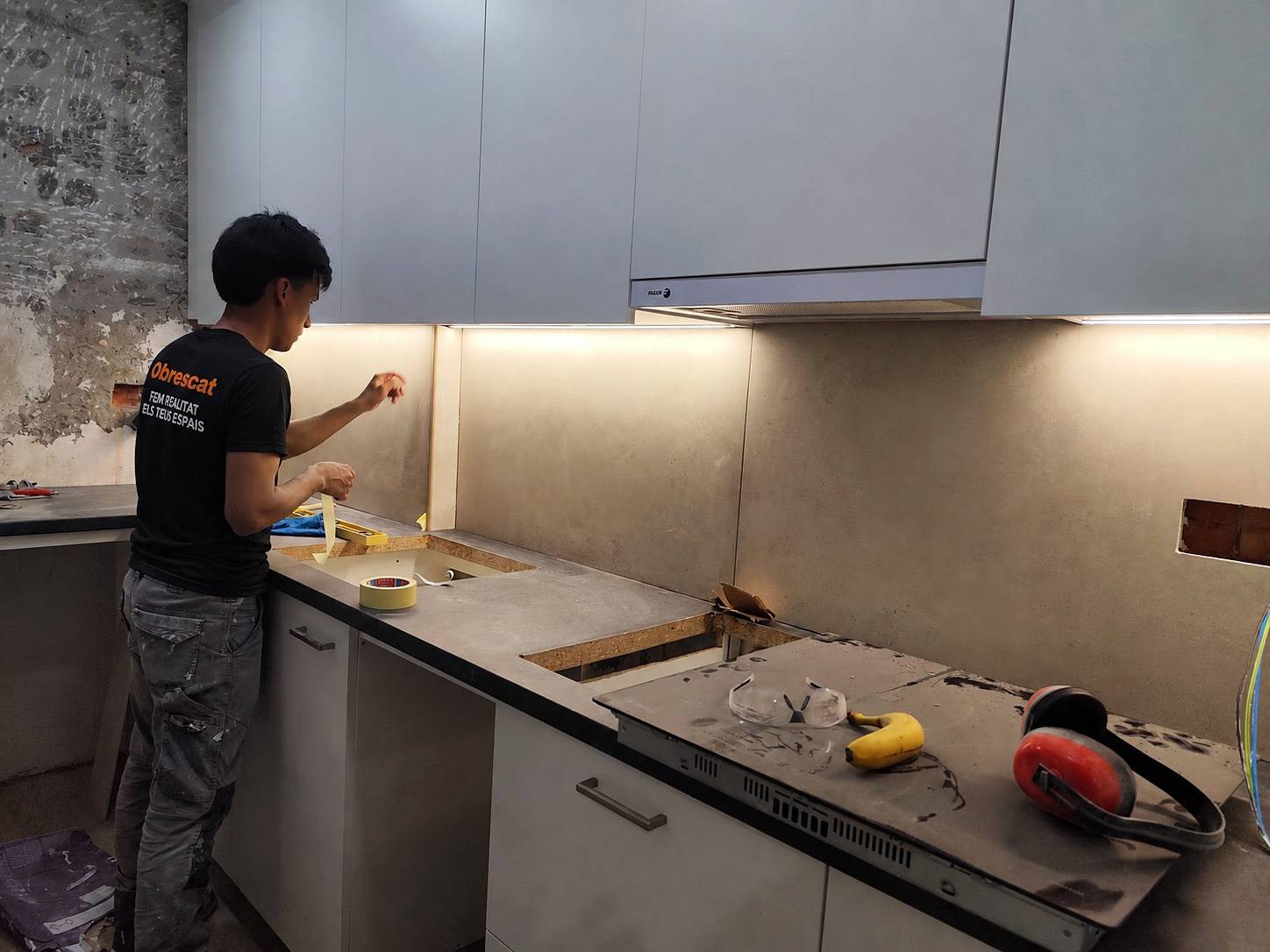 Un montador con la camiseta de Obrescat prepara la encimera de una cocina durante el montaje, con la luz bajo los altos encendida